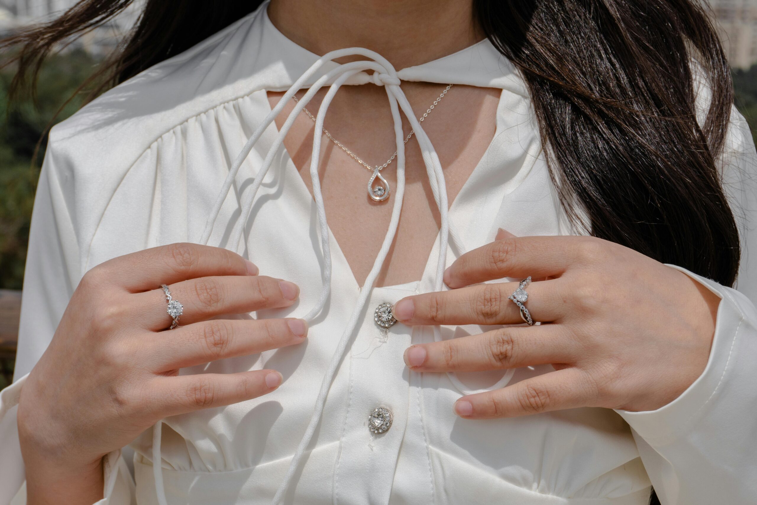 Nahaufnahme einer Frau, die im Freien eleganten Schmuck, darunter Ringe und eine Halskette, mit einer weißen Bluse trägt.